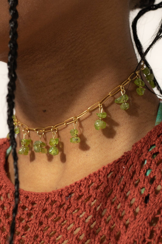 Bijoux ras de cou de couleur dorée avec perles tombantes de couleur verte sur une femme de peau mate avec un top de couleur rouille en crochet