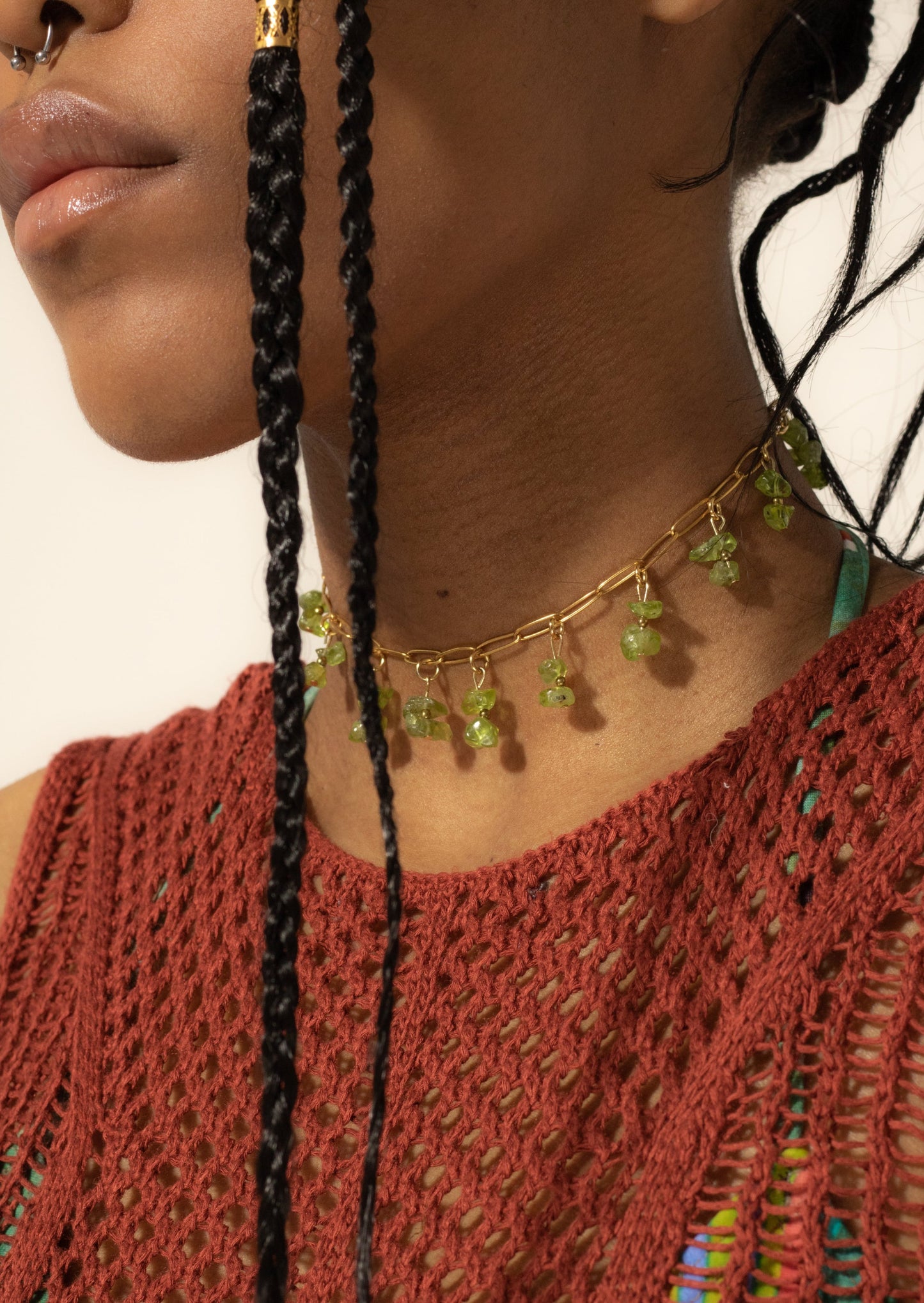 Bijoux ras de cou de couleur dorée avec perles tombantes de couleur verte sur une femme de peau mate avec un top de couleur rouille en crochet