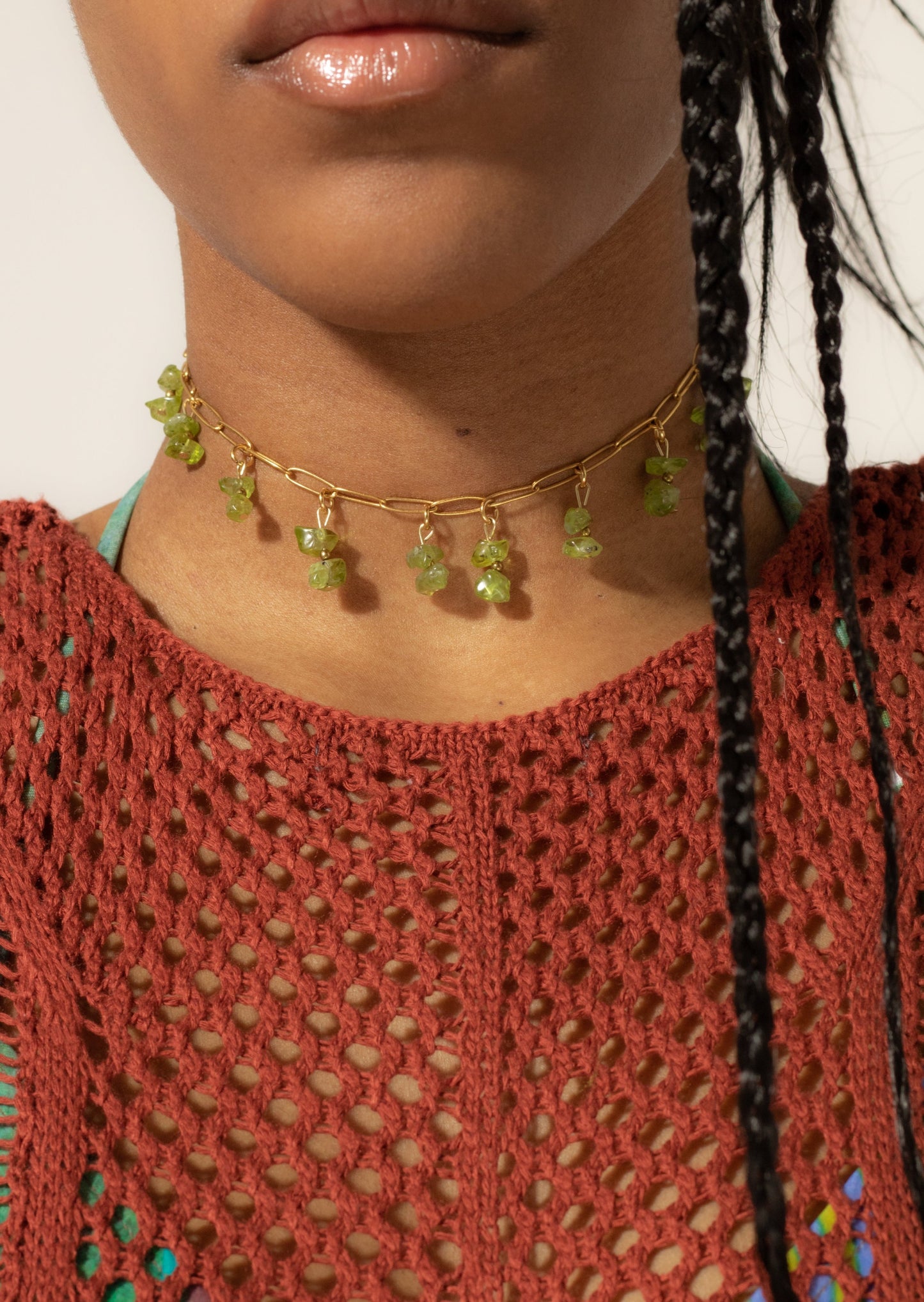 Bijoux ras de cou de couleur dorée avec perles tombantes de couleur verte sur une femme de peau mate avec un top de couleur rouille en crochet